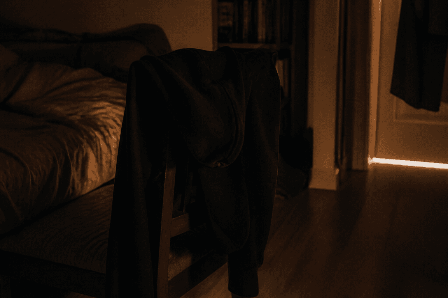 A dark bedroom at night, black clothing over a wooden chair, a strip of streetlight under the door